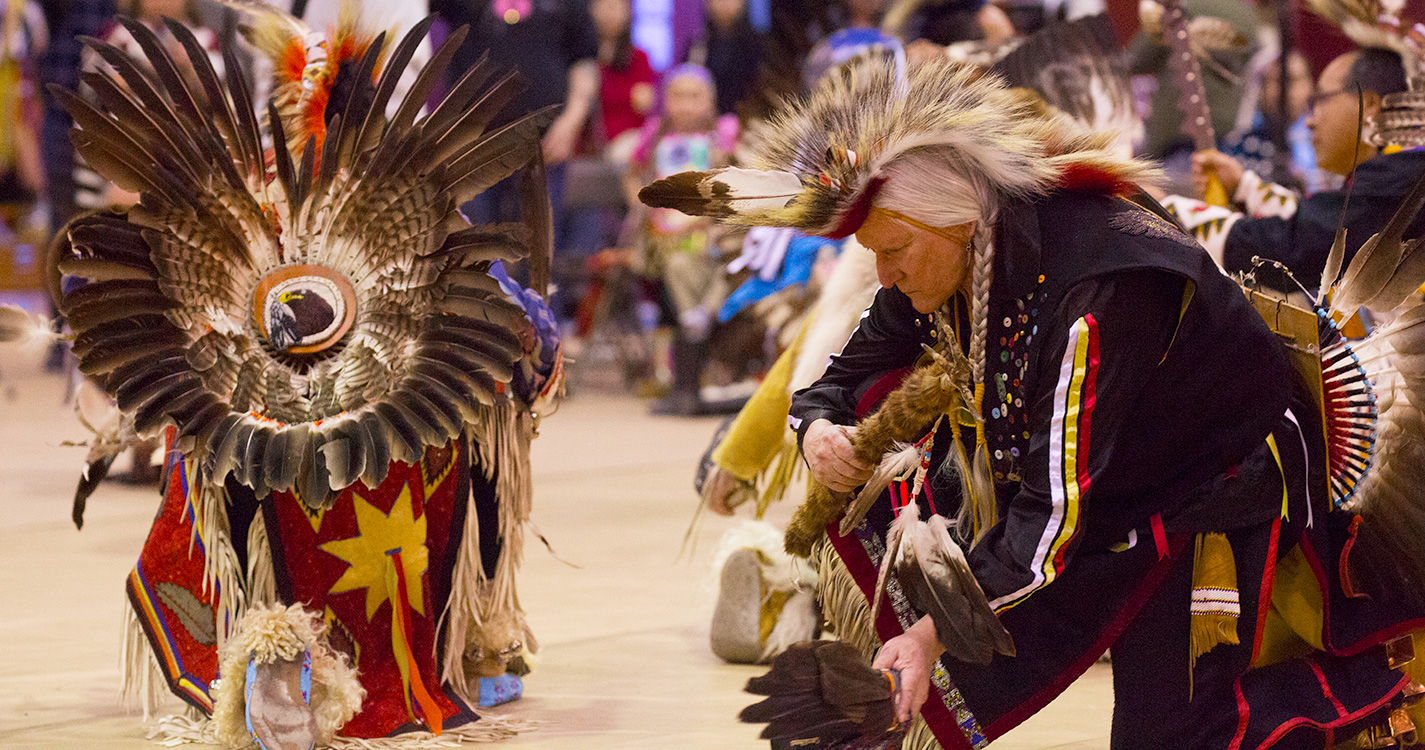 Two men wearing regalia knees during the Social Pow wow.