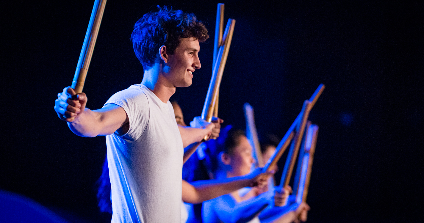 man with arms extended holing drum-stick like objects while dancing in the luau