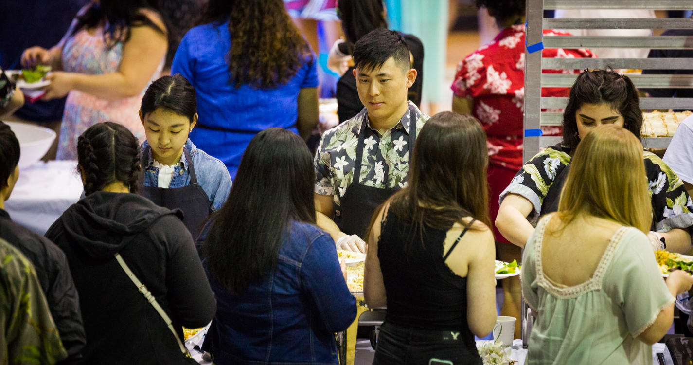 students serve a meal that they cooked to a line of people in a gymnasium