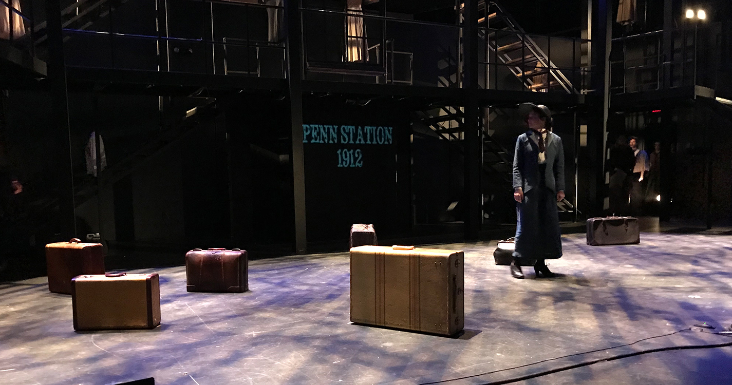 A woman stands onstage surrounded by suitcases in low blue light