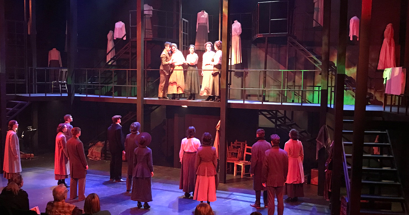 A crowd bathed in red light looks up at five people standing on a balcony in white light