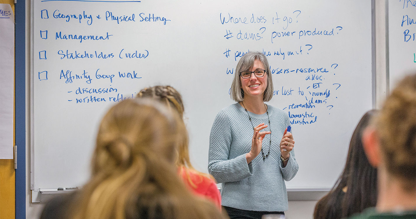 Professor Karen Arabas stands before a classroom with a whiteboard with Overview Colorado River written on it.