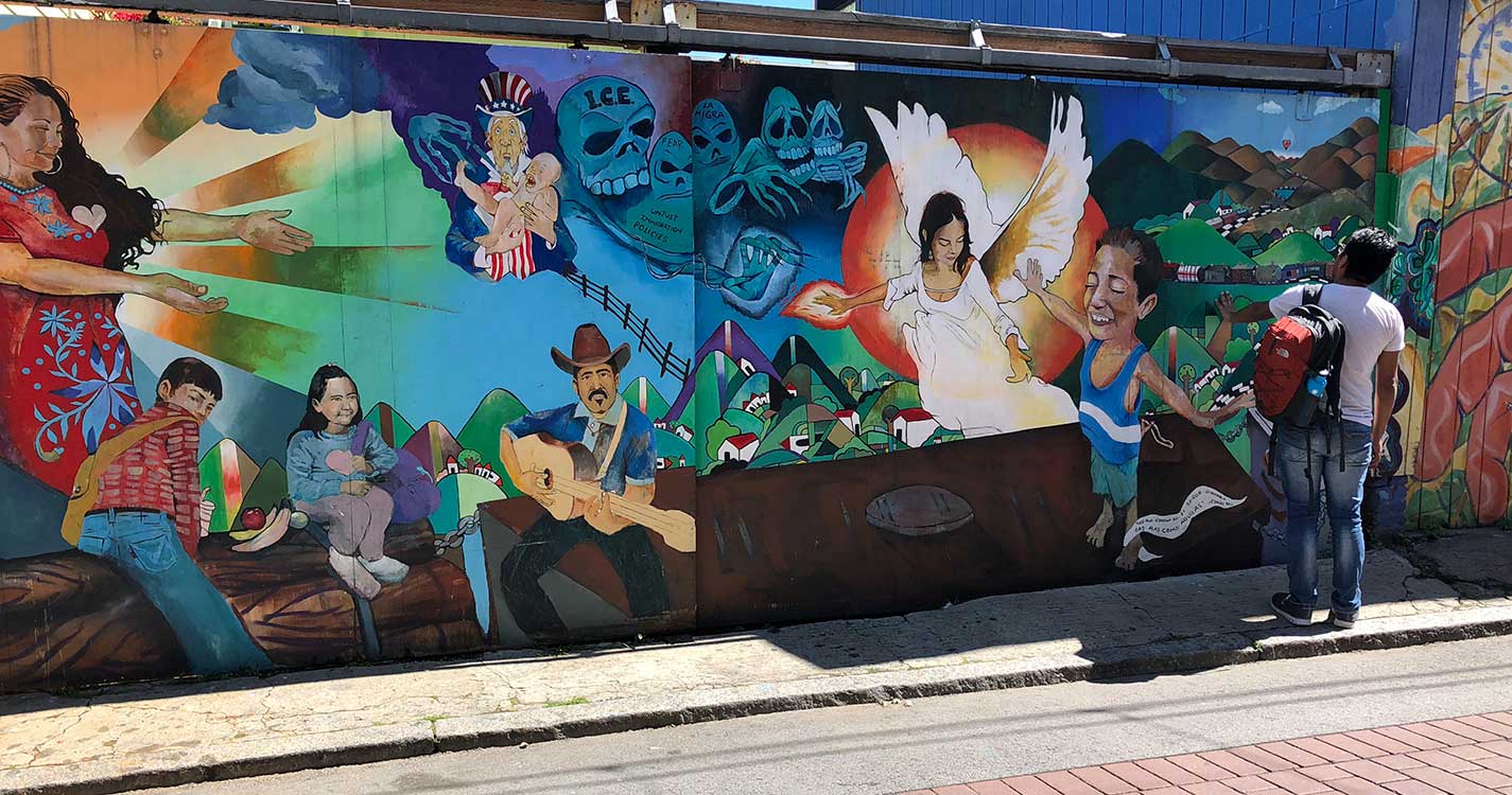 A person with back to camera stands looking at a colorful mural that depicts immigrants' journey to the U.S.A.