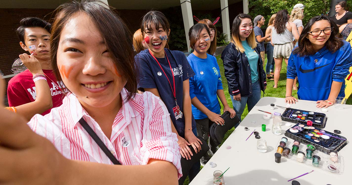 ASP students gather around a student who is face painting the photographer of the photo.