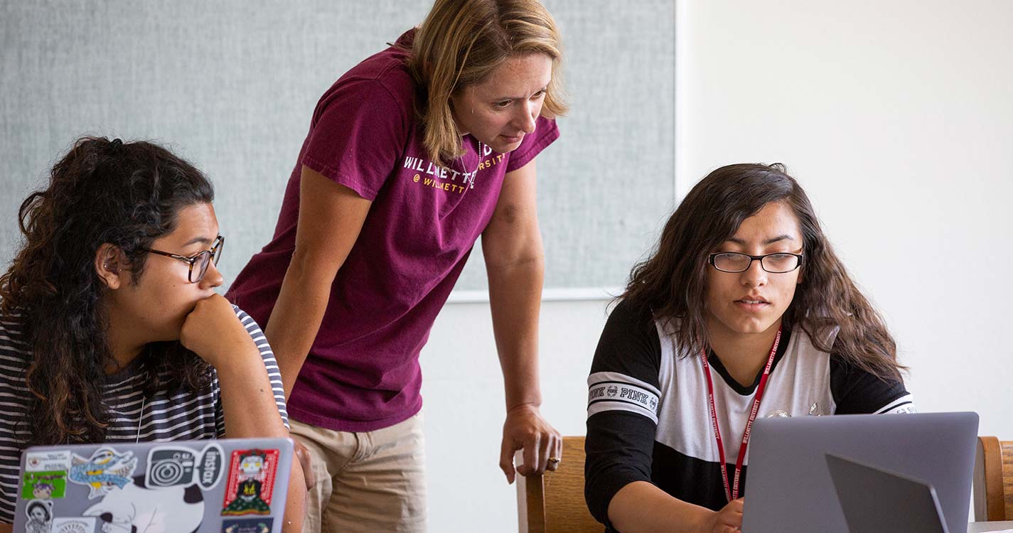 A teacher helps two students who sit at desks with open laptops