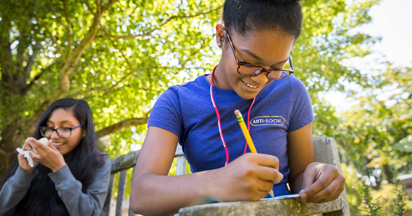 A student sitting under a tree writes in a notebook