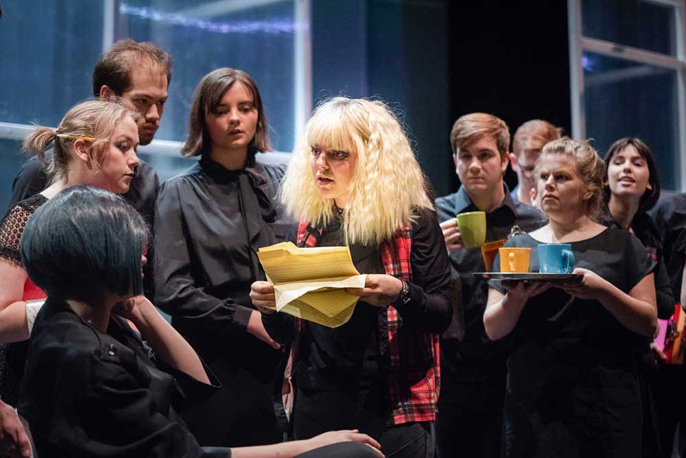 A woman with blonde hair and dark eye makeup stands holding pages ripped from a yellow pad