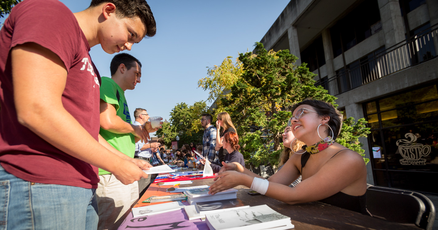 A student sitting at an expo table speaks to a student standing across the table