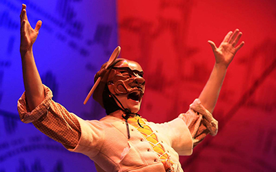 Character wearing wooden mask with hands outstretched in a Y before a blue and red backdrop