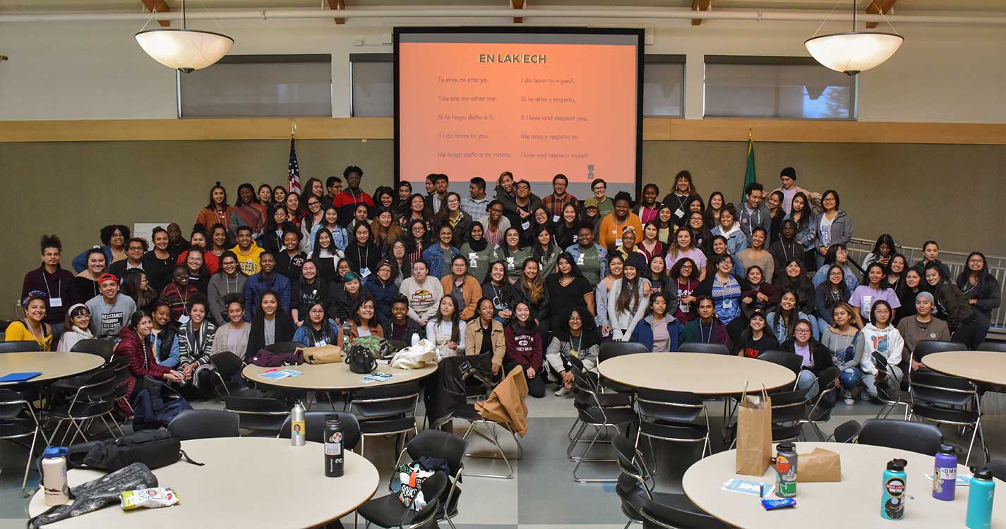 A large group of students, roughly 100, stand for a photo in a conference room