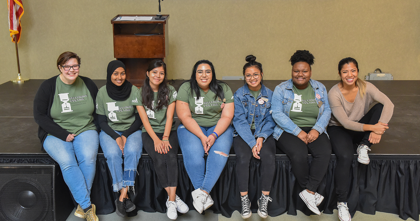 Seven students sit on the edge of a low stage while smiling