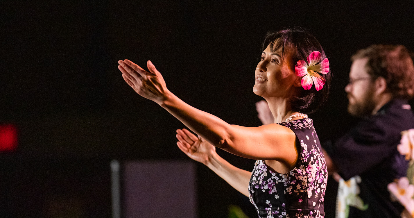 Professor with flower in hair dances onstage at lū’au