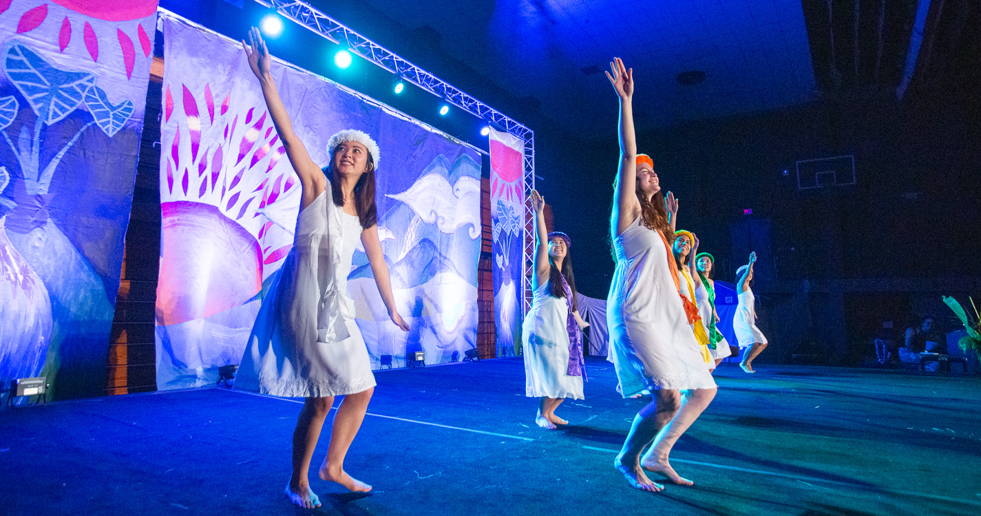 wide view of three dancers onstage in white dresses with arms extended