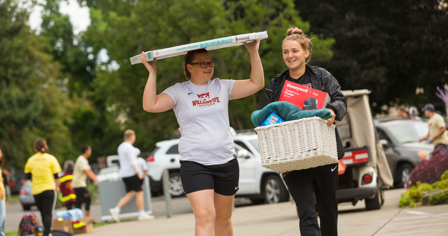 A student carries a basket of clothing while another balances a bulletin board on their head