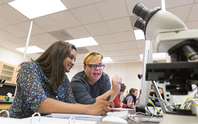 student and professor crouch over a computer screen next to a microscope