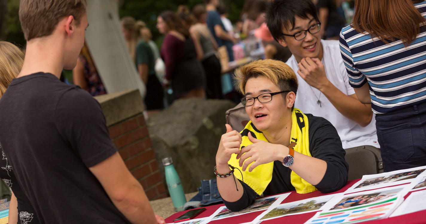 A smiling student seated at a table outside helps a standing student sign up for a club