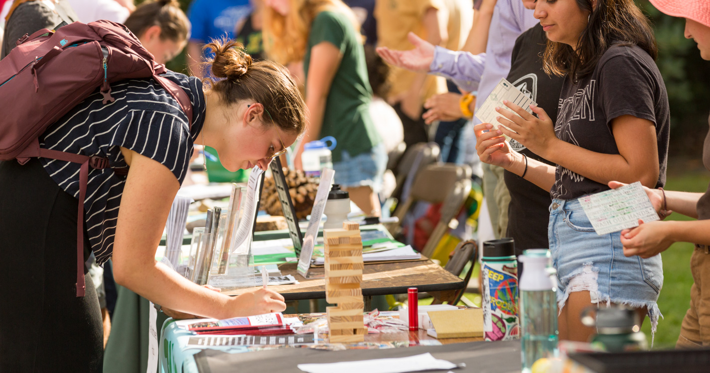 A student signs up on a list at a table in a line of tables along the Mill Stream