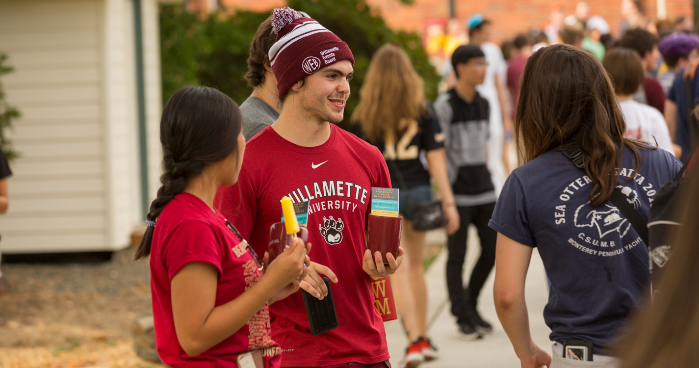 Two students wearing cardinal Willamette swag converse. One holds a Possible. 