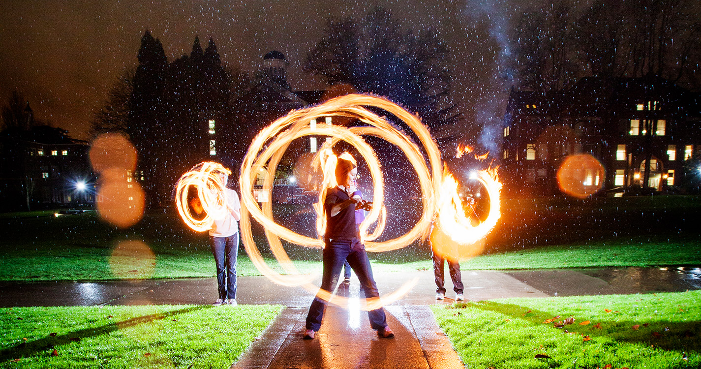 three students twirl flaming poi in a time-lapse photo so the fire forms circles around them