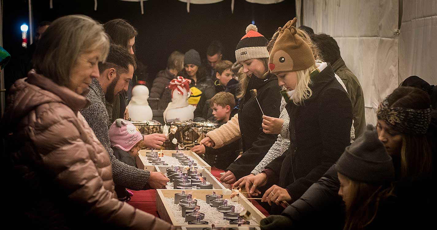 People lined up along table roast s'mores over small fires