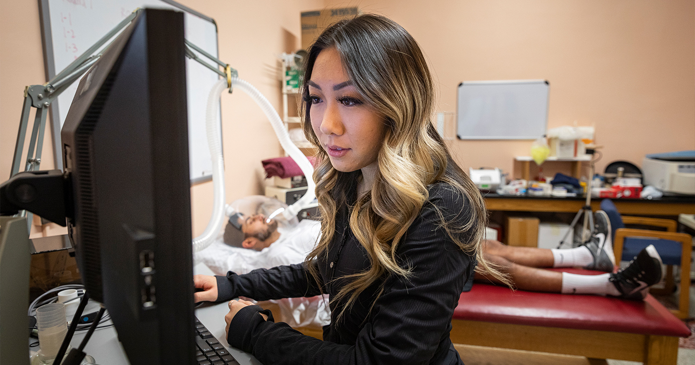 Katie Lee at a computer in a research lab with a student lying on a bed with a ventilator on