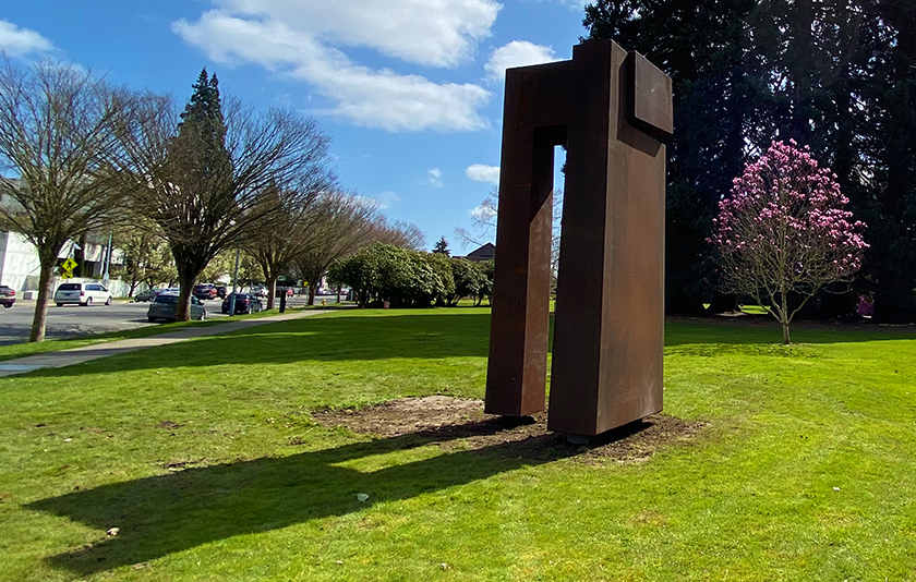 Sculpture of two columns with a cross beam in front of a small pink flowering tree and towering Star Trees