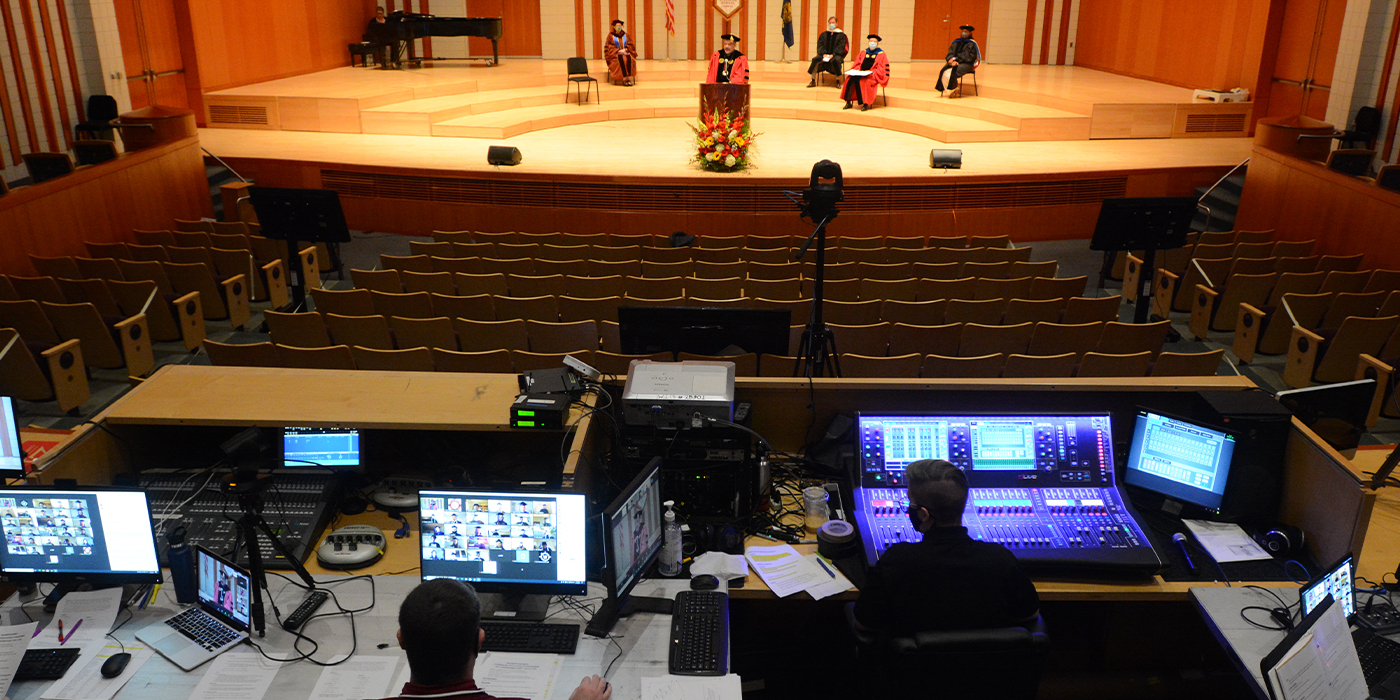 President Thorsett speaks from a podium while technitions work behind a collection of computer monitors to broadcast the interactive event.