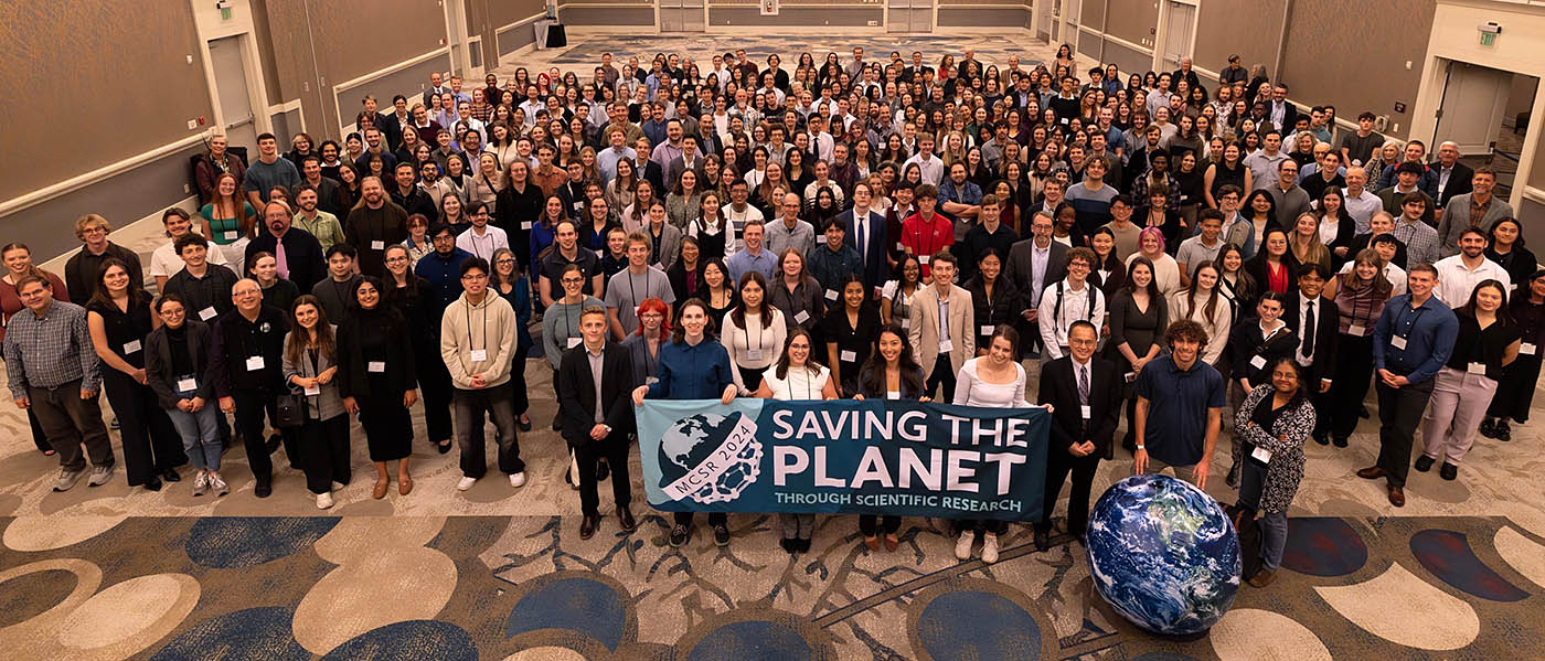 Connor Everetts BS’26 and a large group of people with a "Saving the Planet Through Scientific Research" banner