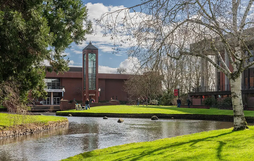 View of the clock tower, the Mill stream, green grass, and trees under a partly cloudy sky.