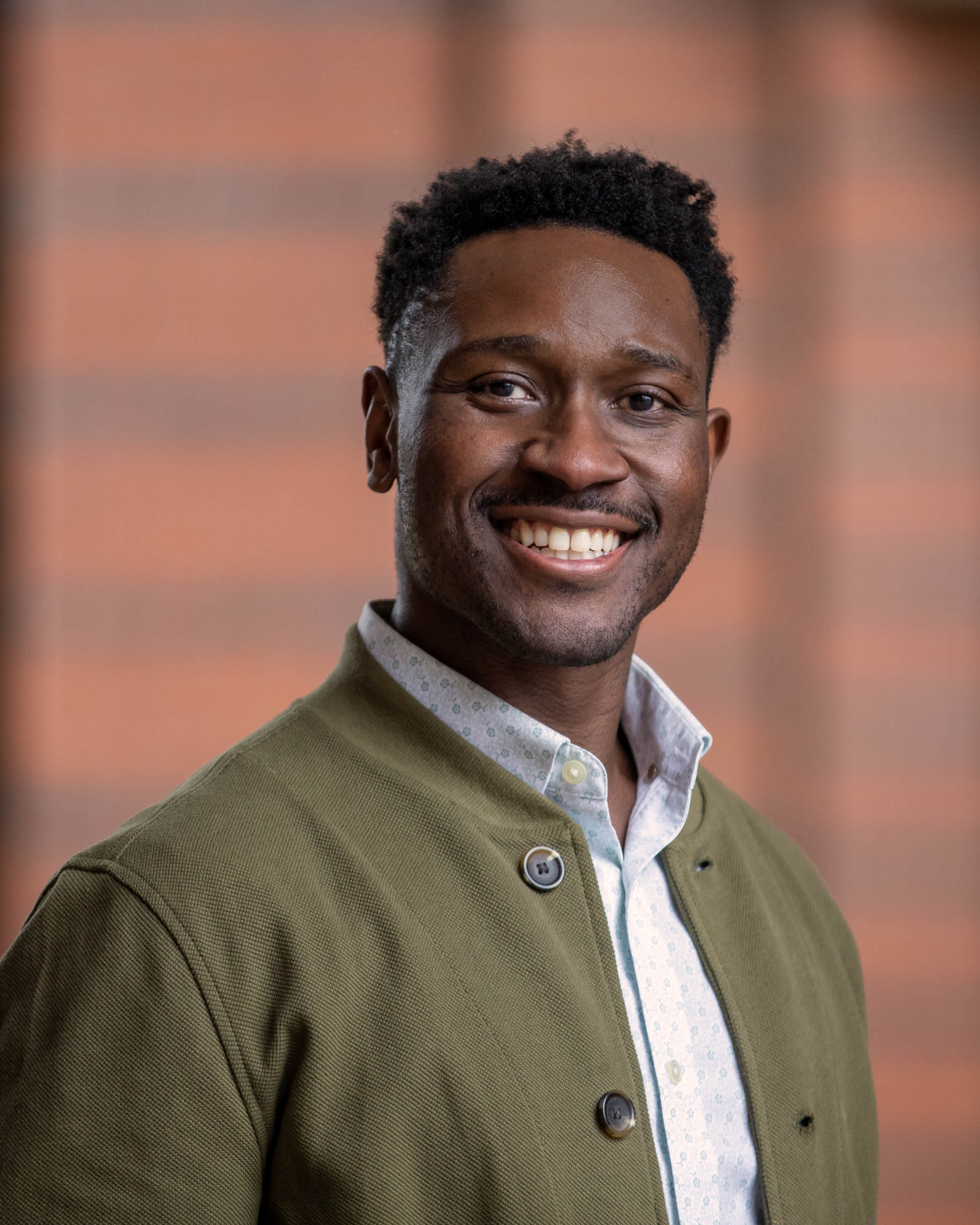 Image of a black man from the waist up, with a mustache, olive green jacket and white button down shirt with a blue pattern