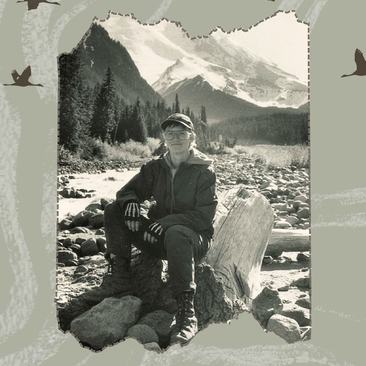 Image of person sitting on a log, in front of tree lined stream, with mountains in the distance